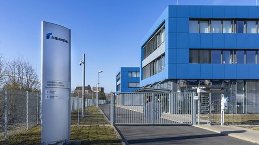 Zukunft zu einem Firmengelände, links eine Säule mit Firmenlogo, hinter einem Tor mehrere blaue Gebäude