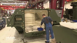 Ein Arbeiter in Latzhose in einer Fabrikhalle arbeitet an einer dunkelgrünen Fahrzeug-Karosserie