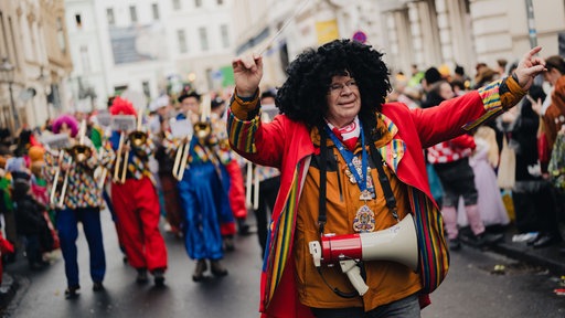 Ein Mann mit Afro-Perücke und Dirigentenstab steht vor einer Blaskapelle.