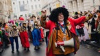 Ein Mann mit Afro-Perücke und Dirigentenstab steht vor einer Blaskapelle.
