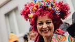 Eine Frau mit roter Perücke und bunt bemaltem Gesicht lacht in die Kamera.