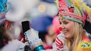 Eine Frau fotografiert eine andere Frau mit bunter Narrenkappe, die sich einen Lutscher vors Gesicht hält.