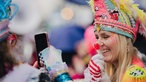 Eine Frau fotografiert eine andere Frau mit bunter Narrenkappe, die sich einen Lutscher vors Gesicht hält.