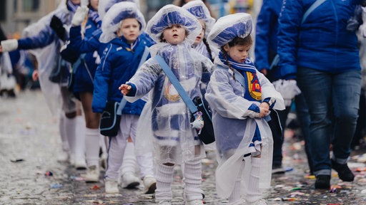 Kinder in blau-weißer Gardeuniform mit durchsichtigen Regencapes stehen in einer Reihe.