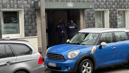 Zwei Polizisten an einer Wohnungstür, im Vordergrund ein blau-weisser Mini-Cooper