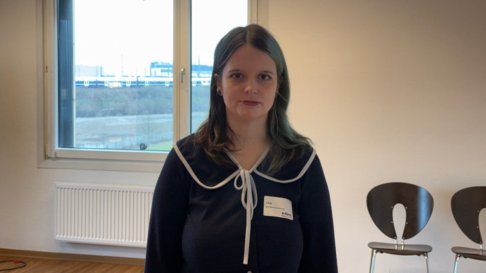 Auf dem Bild sieht man eine Frau mit einem schwarzen Oberteil, im Hintergund ist ein Fenster und zwei Stühle zu sehen. Die Frau heißt Lina.