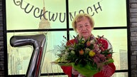 Eine Frau in rotem Blazer mit Blumenstrauß vor Luftballons mit der Zahl 70