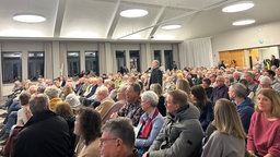 Viele Menschen sitzen in einem Saal, ein Mann steht und spricht in Mikrophon 