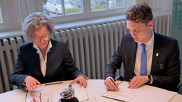 Ina Scharrenbach und Daniel Flemm sitzen an einem Tisch und unterschreiben jeweils ein Schriftstück.