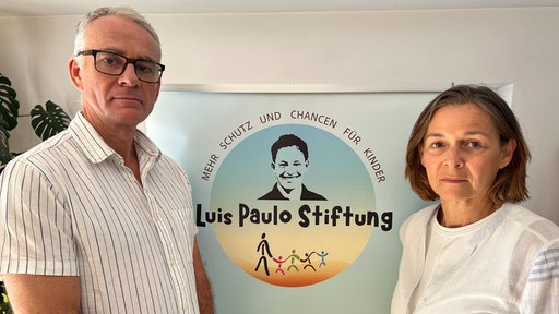 Eine Frau und ein Mann stehen vor einer Plakatwand mit der Aufschrift "Luis Paulo Stiftung" und blicken ernst.
