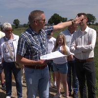 Gruppe Menschen bei einem Feld, Mann vorne hält Zettel in der Hand und zeigt in die Ferne