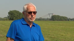 Ein Mann mit grauen Haaren, Sonnenbrille und blauem Poloshirt vor einer großen Wiese