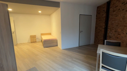 In einem Raum mit gemauerter Wand steht ein unbezogenes Bett sowie ein Tisch mit Stühle, ansonsten ist der Raum leer