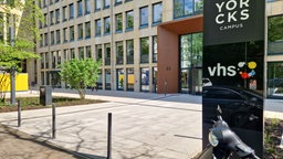 Im linken Teil des Bildes sieht mein ein braunes Gebäude, rechts vorne ein schwarzes Schild mit der Aufschrift VHS.