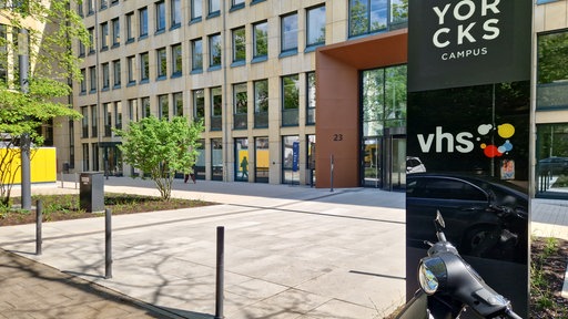 Im linken Teil des Bildes sieht mein ein braunes Gebäude, rechts vorne ein schwarzes Schild mit der Aufschrift VHS.