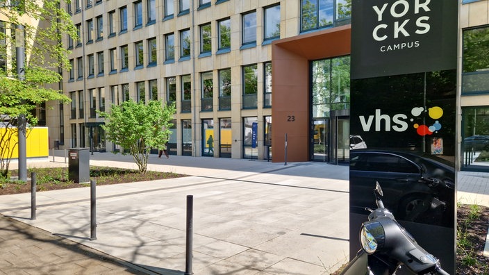 Im linken Teil des Bildes sieht mein ein braunes Gebäude, rechts vorne ein schwarzes Schild mit der Aufschrift VHS.
