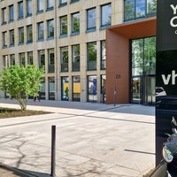Im linken Teil des Bildes sieht mein ein braunes Gebäude, rechts vorne ein schwarzes Schild mit der Aufschrift VHS.