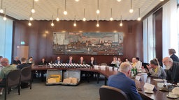 Saal mit halbrundem Tisch, auf vielen Stühlen Menschen, Ordner auf dem Tisch