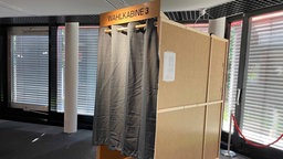 In einem Raum steht eine Wahlkabine mit zugezogenem Vorhang