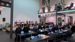 In einem Raum mit hohen Decken sitzen Menschen auf einem Podium und weitere Menschen an Stühlen mit Namensschildern