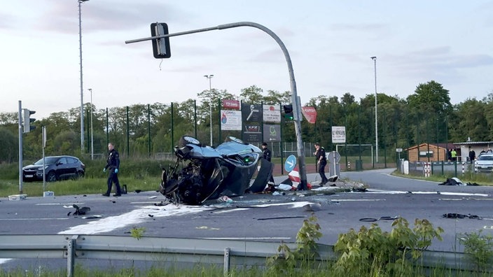 Auf dem Bild ist eine Kreuzung zu sehen. Mittig im Bild ist ein Autowrack, dass auf der Seite liegt. Im Hintergrund ist ein Sportplatz zu sehen.