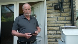 Ein älterer Mann steht mit einem Handy in der Hand neben dem Briefkasten vor seiner Haustür.