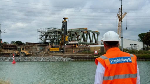 Mann mit Warnweste auf der "Projektleitung DB" seht, betrachtet Großbaustelle.