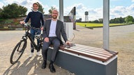 CDO Denez Kücük steht mit einem E-Bike neben einer der Smart-Bänke. Stadtdirektor Sebastian Kopietz sitzt auf der Bank.