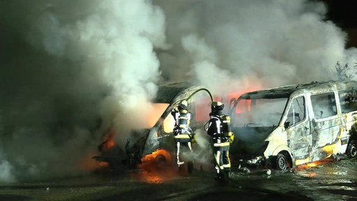 In Bochum sind zwei Transporter ausgebrannt. Zwei Feuerwehrleute löschen die Fahrzeuge, die in Vollbrand stehen.