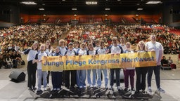 Eine Gruppe junger Menschen steht auf einer Bühne. Sie halten ein Banner mit der Aufschrift "Junge Pflege Kongress Nordwest". Im Hintergrund sitzen Menschen im Publikum.