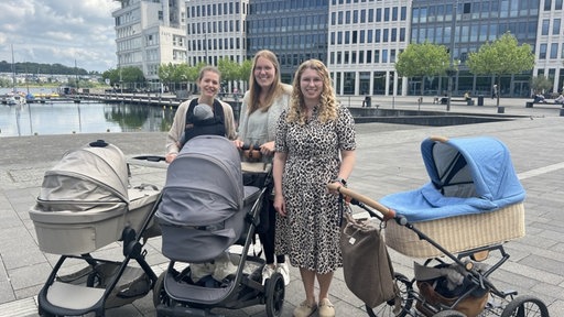Kim Kloth und ihre zwei Freundinnen stehen gemeinsam mit drei Kinderwägen in einem Park.