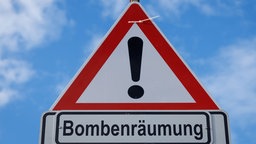 Auf dem Foto ist ein Schild mit einem rot umrahmten Ausrufezeichen und dem Schriftzug "Bombenräumung" darunter.