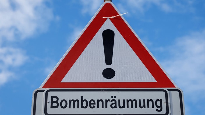 Auf dem Foto ist ein Schild mit einem rot umrahmten Ausrufezeichen und dem Schriftzug "Bombenräumung" darunter.