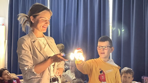 Frau steht mit Taschenlampe in Klassenraum. Junge hält Hand über Taschenlampe.