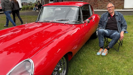 Ein Mann sitzt auf einem Campingstuhl neben seinem roten Jaguar E-Type.