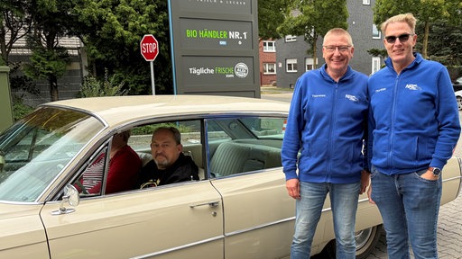Zwei Männer in blauen Sweatjacken stehen vor einem Cadillac an der Einfahrt zum Oldtimer Treffen.