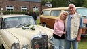 Ein älteres Paar steht Arm in Arm neben ihrem Mercedes 220, der hochzeitlich geschmückt ist.