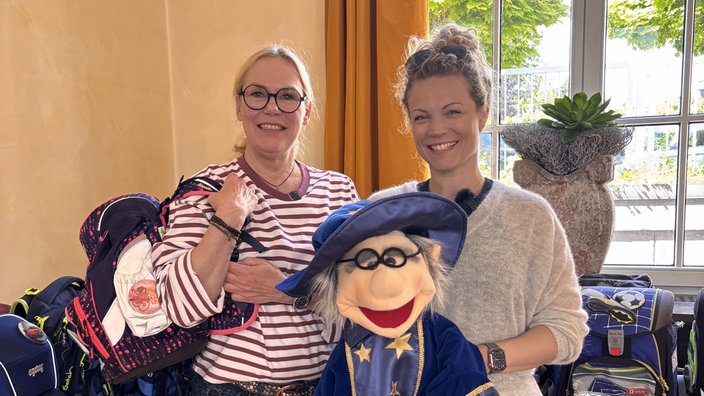 Jessica Helmke und Claudia Belinski mit dem Wunschzauberer-Maskottchen und einem Tornister