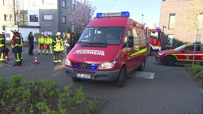Mehrere Fahrzeuge der Feuerwehr stehen auf einem Parkplatz zwischen Gebäuden, daneben stehen Feuerwehrkräfte