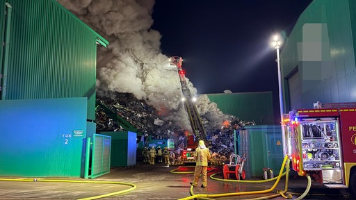 Au dem Schrottplatz in Herne wütet ein Feuer. Feuerwehrmann und Feuerwehrwagen versuchen den Brand zu löschen.