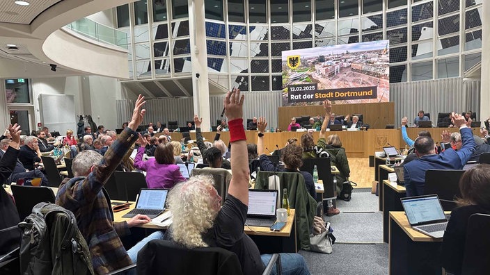 Mehrere Personen sitzen im Halbkreis im Rat der Stadt Dortmund und heben die Hand