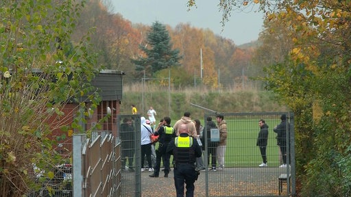 Menschen stehen an einem Fußballplatz, zwei Polizisten kommen gerade dazu.