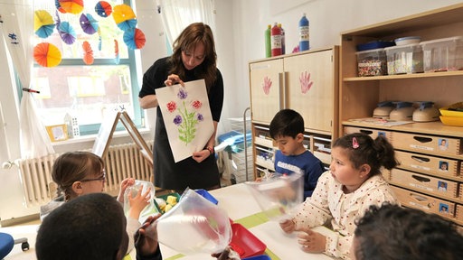 Mehrere Kleinkinder sitzen in der Kita an einem Tisch, vor ihnen steht eine Pflegerin und zeigt ihnen ein Bild