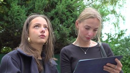 Auf dem Foto sind zwei junge Frauen. Eine schaut interessiert noch vorne, die andere notiert etwas auf ihrem Tablet.