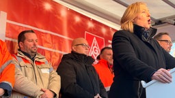 Bärbel Bas spricht auf einem Podium, hinter ihr stehen Männer in einer Reihe.  