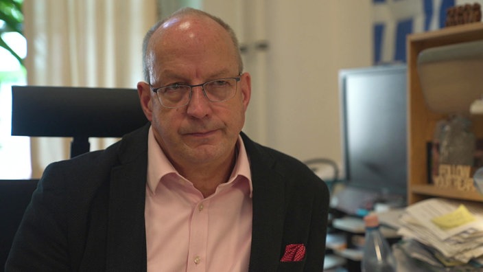 Schulleiter Christof Hearing guckt ernst in die Kamera. Er trägt eine Brille, ein rosa Hemd und ein schwarzes Sakko darüber.