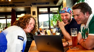 Drei junge Männer sitzen in einer Gaststätte an einem Tisch und blicken auf einen Laptop. Im Hintergrund sind Pflanzen, Fenster und weitere Gäste zu sehen.