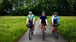 Drei Radfahrer fahren nebeneinander auf einem schmalen Asphaltweg durch eine grüne Wiesenlandschaft.