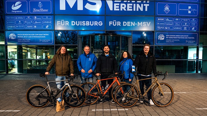 Fünf Personen stehen mit drei Fahrrädern vor dem Eingangsbereich eines modernen Gebäudes mit großer blauer Glasfassade. Das Stadion vom MSV Duisburg. Im Hintergrund sind Schriftzüge, Logos und beleuchtete Fenster zu sehen.