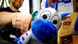 Ein blau-weißes Plüschzebra wird von einer Person in die Kamera gehalten. Im Hintergrund sind unscharf weitere Personen und der Innenraum einer Gaststätte zu sehen.